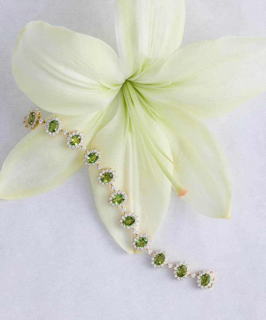 Gold bracelet with green gems and diamonds draped over a white lily.